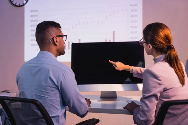 Businesswoman in glasses pointing with finger at computer monitor with blank screen near coworker in office — Stock Photo