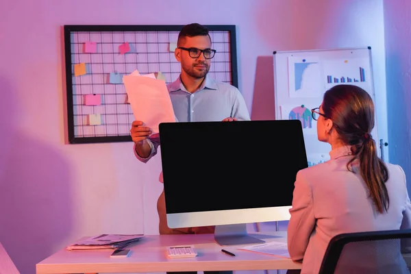 Donna d'affari seduta vicino al monitor del computer con schermo bianco e guardando collega con documenti — Foto stock