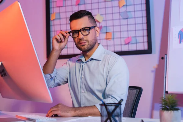 Uomo d'affari che regola gli occhiali vicino alla tastiera del computer e monitor — Foto stock