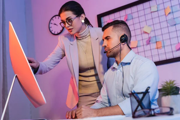 Operatore in auricolare guardando monitor di computer vicino a donna d'affari in ufficio — Foto stock