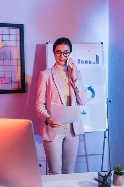 Zufriedene Geschäftsfrau spricht auf dem Smartphone, während sie Dokumente in der Nähe von Flipchart betrachtet — Stockfoto