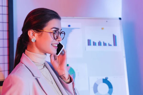 Alegre mujer de negocios hablando en teléfono inteligente cerca de rotafolio con gráficos y gráficos - foto de stock