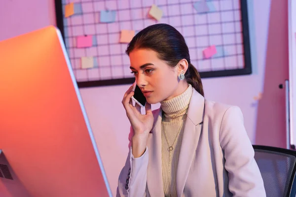 Businesswoman talking on smartphone and looking at computer monitor in office — Stock Photo