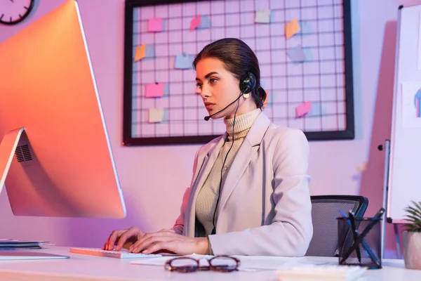 Female operator in headset typing on computer keyboard and looking at monitor — Stock Photo