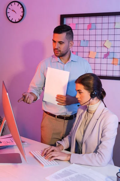 Empresário apontando com o dedo para monitor de computador perto de operador feminino no fone de ouvido — Fotografia de Stock