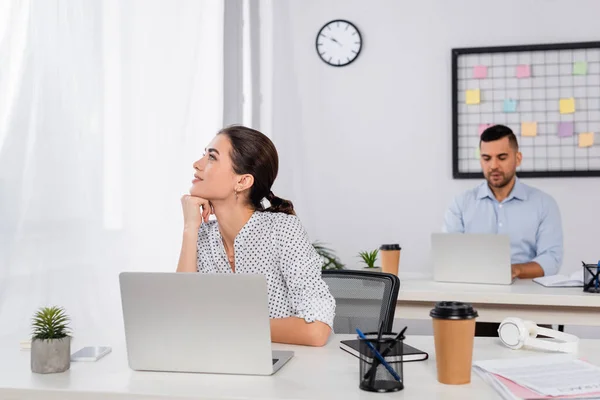 Verträumte Geschäftsfrau schaut in der Nähe von Laptop auf Schreibtisch und Mitarbeiter auf verschwommenem Hintergrund weg — Stockfoto