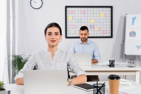 Весела бізнес-леді посміхається біля ноутбука на столі і колега на розмитому фоні — стокове фото