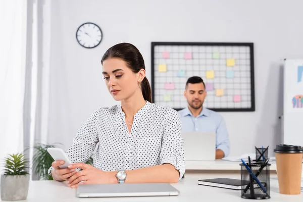 Empresária usando smartphone perto de laptop na mesa e colega de trabalho em fundo embaçado — Fotografia de Stock