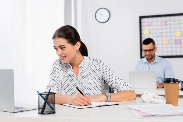 Sonriente mujer de negocios escribiendo en cuaderno cerca de hombre de negocios sobre fondo borroso - foto de stock