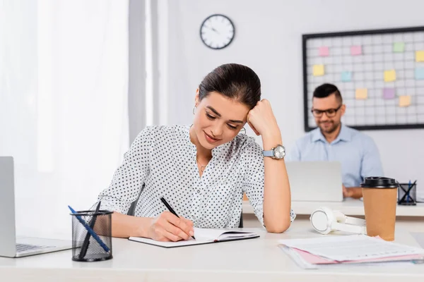 Alegre empresaria escribiendo en cuaderno cerca de hombre de negocios sobre fondo borroso - foto de stock