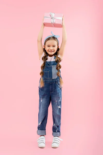 Full length of happy kid smiling and holding present above head on pink — Stock Photo