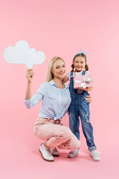 Comprimento total da mãe alegre segurando bolha pensamento enquanto criança de pé com presente no rosa — Fotografia de Stock