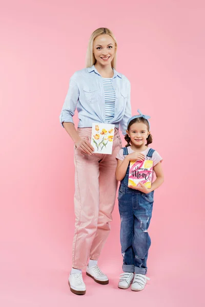 Longitud completa de alegre madre e hija sosteniendo tarjetas de felicitación en rosa - foto de stock