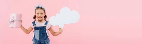 Zufriedenes Kind lächelt und hält Gedankenblase und Geschenk isoliert auf rosa, Banner — Stockfoto