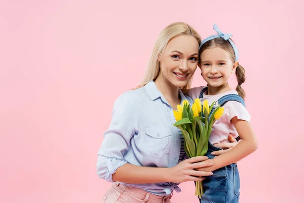 Felice madre che tiene tulipani gialli e abbraccia figlia isolata su rosa — Foto stock