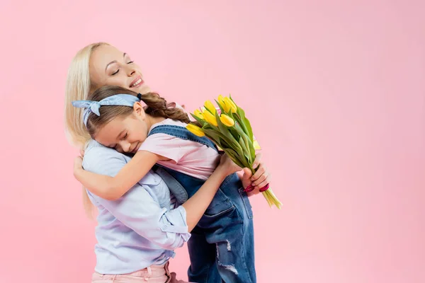 Joyeuse mère tenant des tulipes jaunes et embrassant fille isolée sur rose — Photo de stock