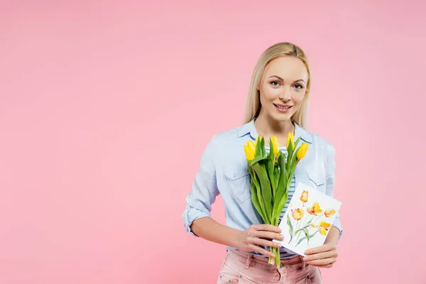 Mujer feliz sosteniendo tulipanes y tarjeta de felicitación aislado en rosa - foto de stock
