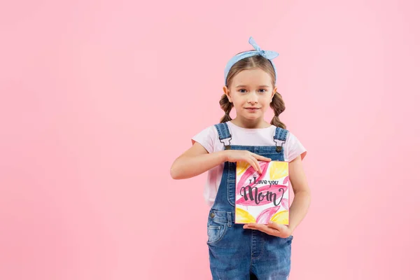 Feliz niño sosteniendo tarjeta de felicitación con te amo mamá letras aisladas en rosa - foto de stock