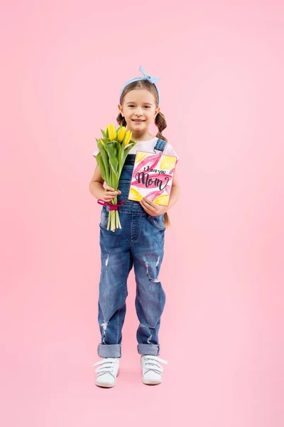 Piena lunghezza del bambino felice che tiene tulipani e biglietto di auguri con ti amo mamma lettering su rosa — Foto stock