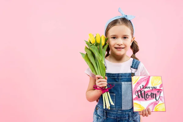 Alegre niño sosteniendo tulipanes y tarjeta de felicitación con te amo mamá letras aisladas en rosa - foto de stock