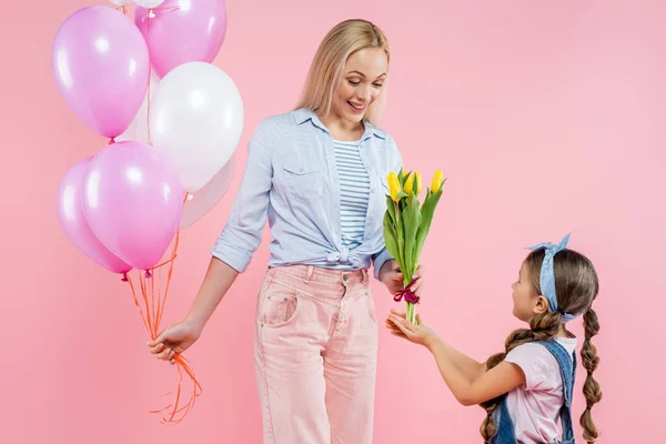 Criança dando tulipas para mãe feliz de pé com balões isolados em rosa — Fotografia de Stock