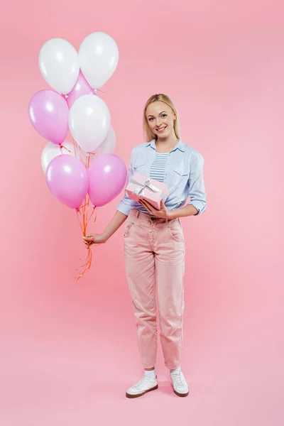 Pleine longueur de femme heureuse tenant des ballons et boîte cadeau sur rose — Photo de stock