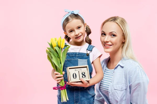 Happy kid holding tulips and wooden cubes with 8 march lettering near mother isolated on pink — Stock Photo