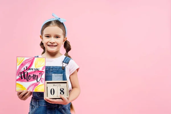 Glückliches Kind hält Grußkarte mit i love you Mama Schriftzug und Holzwürfel mit 8 Marsch isoliert auf rosa — Stockfoto