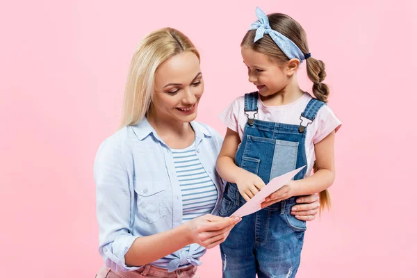 Niño feliz sosteniendo la tarjeta de felicitación cerca de la madre aislada en rosa - foto de stock