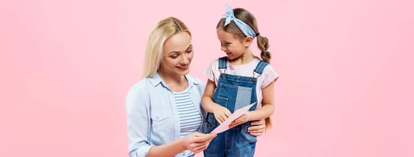 Felice bambino in possesso di biglietto di auguri vicino alla madre isolato su rosa, banner — Foto stock