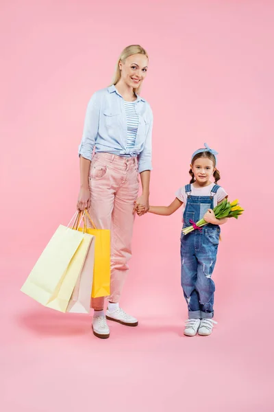 Pleine longueur de mère heureuse avec des sacs à provisions et fille avec des tulipes sur rose — Photo de stock