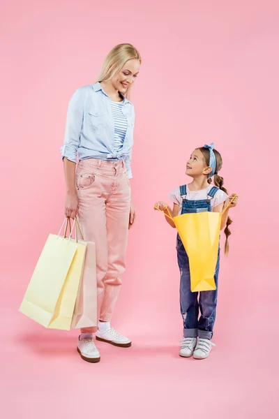 Веселые мать и дочь в полный рост смотрят друг на друга, держа в руках розовые сумки с покупками — стоковое фото