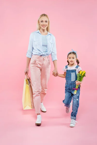Tutta la lunghezza della madre con borse della spesa e figlia con tulipani che camminano sul rosa — Foto stock