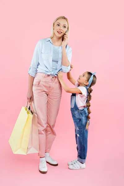 Longitud completa de la madre feliz sosteniendo bolsas de compras y hablando en el teléfono inteligente cerca de la hija en rosa - foto de stock