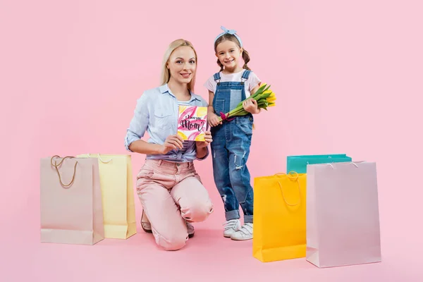 Longitud completa de la madre que sostiene la tarjeta de felicitación cerca del niño con flores y bolsas de compras en rosa - foto de stock