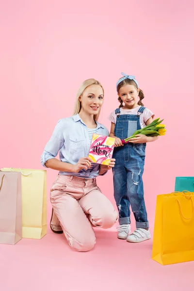 Feliz madre sosteniendo la tarjeta de felicitación cerca del niño con flores y bolsas de compras en rosa - foto de stock