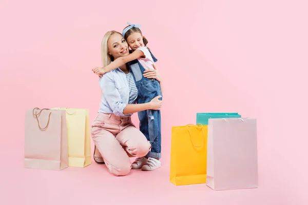 Pleine longueur de fille heureuse étreignant mère souriante près de sacs à provisions sur rose — Photo de stock