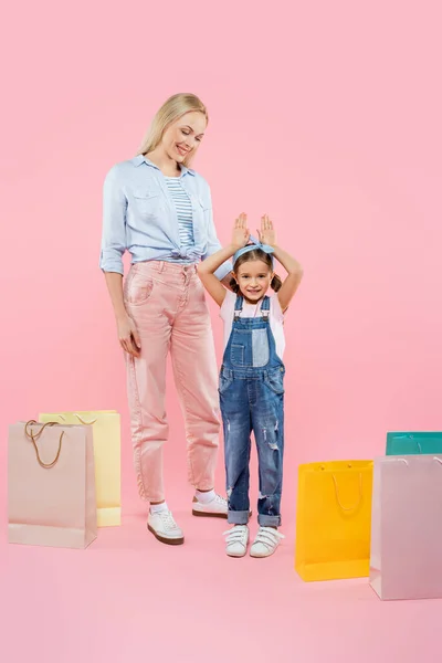 Pleine longueur de mère heureuse regardant enfant avec des oreilles de lapin près de sacs à provisions sur rose — Photo de stock