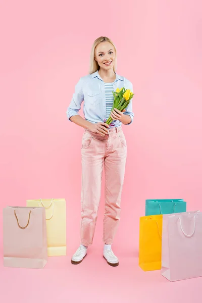 Full length of happy woman holding yellow tulips near shopping bags on pink — Stock Photo