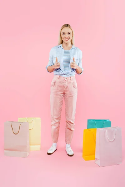 Piena lunghezza della donna felice mostrando pollici in su e in piedi vicino a borse della spesa su rosa — Foto stock