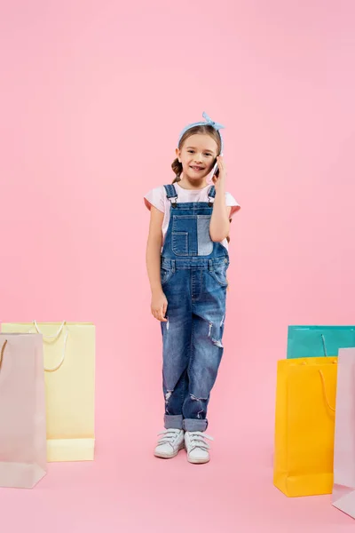 Full length of happy kid talking on smartphone near shopping bags on pink — Stock Photo