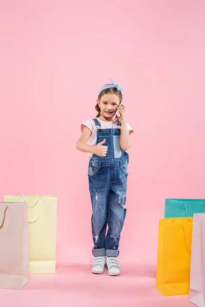 Longitud completa de niño hablando en el teléfono inteligente y mostrando el pulgar hacia arriba cerca de bolsas de compras en rosa - foto de stock