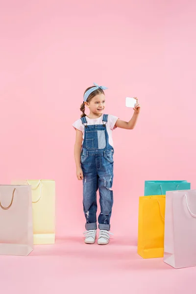 Comprimento total da criança alegre tomando selfie no smartphone perto de sacos de compras em rosa — Fotografia de Stock