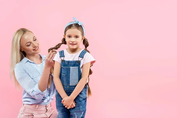 Mãe feliz trançando o cabelo da filha sorridente isolada no rosa — Fotografia de Stock