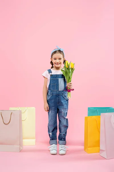 Longitud completa de niño feliz sosteniendo tulipanes cerca de bolsas de compras en rosa - foto de stock