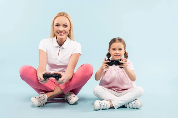 KYIV, UCRANIA - 25 de noviembre de 2020: madre e hija alegres sosteniendo joysticks mientras están sentadas con las piernas cruzadas en azul - foto de stock