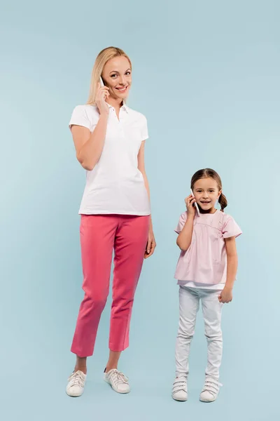 Full length of cheerful mother and daughter talking on smartphones on blue — Stock Photo