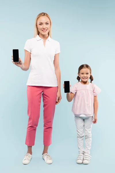 Full length of cheerful mother and daughter holding smartphones with blank screen on blue — Stock Photo