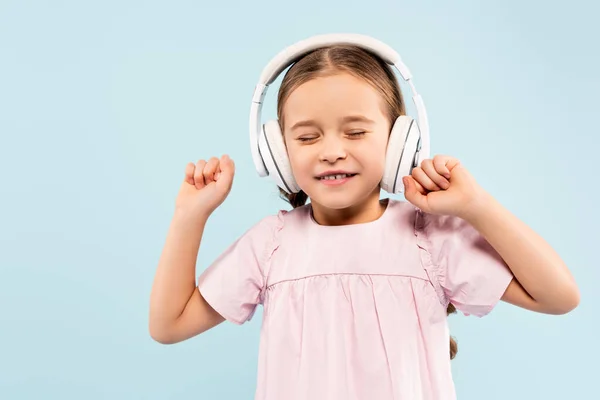 Niño feliz con los ojos cerrados en auriculares inalámbricos sonriendo aislado en azul - foto de stock