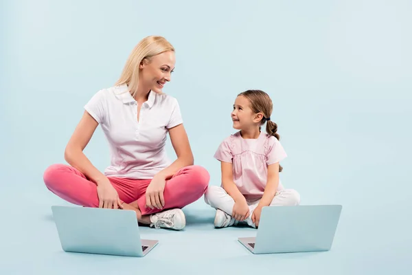 Happy mother and daughter sitting with crossed legs near laptops and looking at each other on blue — Stock Photo